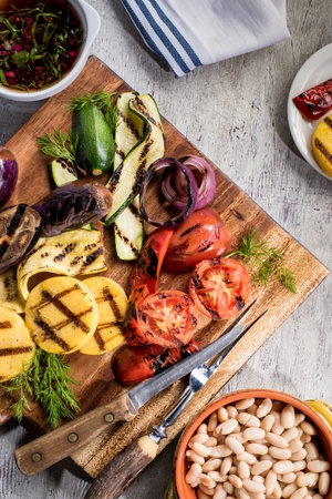 Grilled zucchini, onions, tomatoes and beans on a wooden cutting board with utensilsの写真素材
