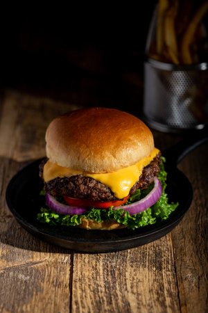 Hot Cheeseburger with melting cheese, lettuce, tomato on a brioche bun with french fries in a conical holder. Dark wood surfaceの写真素材