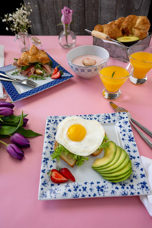 Brunch for two people with a beautiful pink background, Croissants, avocadoes, orange juice, eggs on toast and a parma sandwich. Fresh tulips in the left cornerの写真素材