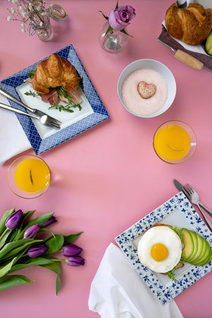 Brunch table setting with eggs, Croissants, avocado, toast orange juice and fresh flowers on a pink background. Area to put message in middleの写真素材