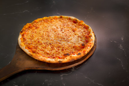 A whole uncut wood fired pizza on a pizza peel sliding onto a black marble counter topの写真素材