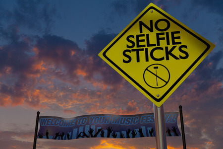 A no selfie sticks sign rises above a banner welcoming guests to a music fetival with a sunset in the background.の写真素材
