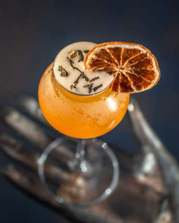 Orange cocktail glass decorated with a slice of dehydrated orange resting on a metal sculpture in the shape of a hand.の写真素材