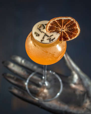 Orange cocktail glass decorated with a slice of dehydrated orange resting on a metal sculpture in the shape of a hand.の写真素材
