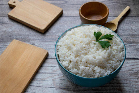 White rice in a green bowl. There are two wooden bowls, a wooden stove, two cutting boards, and a green bowl. All on a rustic gray table.の写真素材