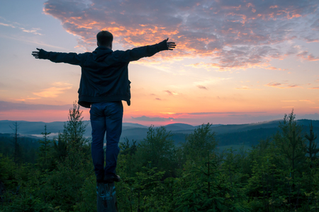 A young guy stands on a log, looks from a height to the mountains with his arms outstretchedの写真素材