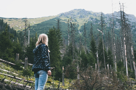Young blonde girl looking at fallen trees in spring forest on background of mountainsの写真素材