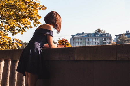 Young girl in blue dress with a bouquet of yellow autumn leaves looking at the autumn cityscapeの写真素材