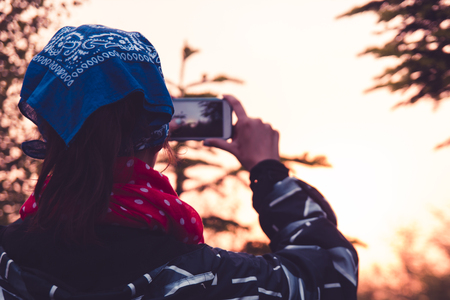 A young girl in a blue shawl stands with her back and takes pictures of nature and mountains at dusk at sunset on a mobile phone.の写真素材