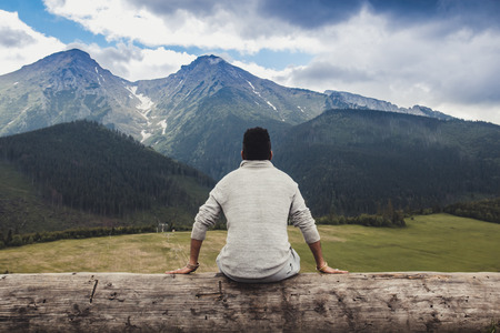Young African Americans guy sitting on a big log and looking at the beautiful panorama of the mountains before the stormの写真素材