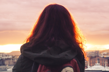 A lonely girl with a backpack stands with her back and looks at the beautiful panorama of the city at sunset, concept of tranquility, travel and serenityの写真素材
