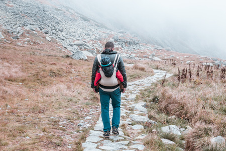A young father carries on the shoulders of a small child, he walks along a stone mountain path in the fogの写真素材