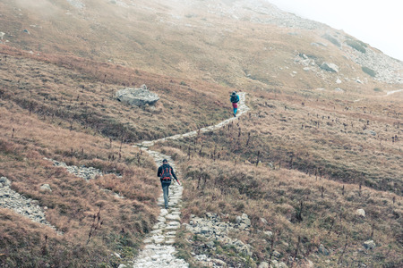 Two travelers hike a stone trail among the mountains and red dry grassの写真素材