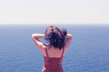 Young brunette girl in red dress looking at blue seascape and enjoying summer vacationの写真素材