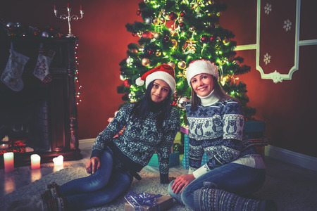 Young female friends in hats Santa Claus near a Christmas tree in the decorated room.の写真素材