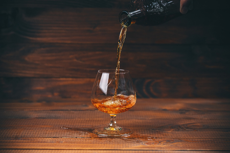Pouring brandy or cognac from the bottle into the glass against wooden backgroundの写真素材