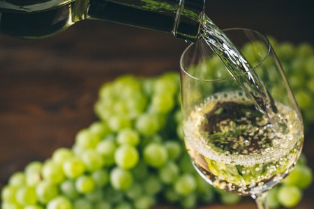 White wine poured into a glass with a bunch of green grapes against wooden backgroundの写真素材