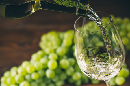 Pouring white wine into a glass with a bunch of green grapes against wooden backgroundの写真素材