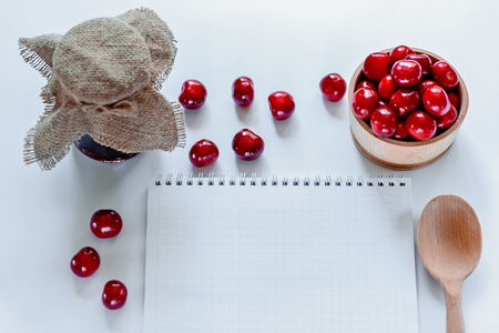 Red ripe cherries with jam jar, wooden spoon and notebook on white background. Flat lay. Food concept.の写真素材