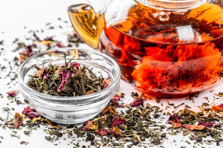 Tea in a glass teapot with a blooming large flower. Teapot with exotic green tea on a white background with scattered dried tea with petals and pieces of fruitの写真素材