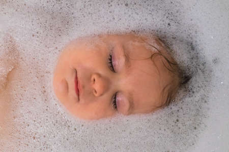 child girl bathes in a bathtub with foam white bath blonde contented face smile beautiful eyes wet...