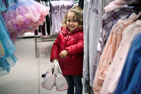 Little Girl in red jacked with Two bags  Looking Curiously in the Store for Salesの写真素材