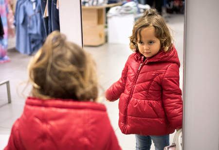 Little Girl in red jacked Looking in the Mirror in the Storeの写真素材