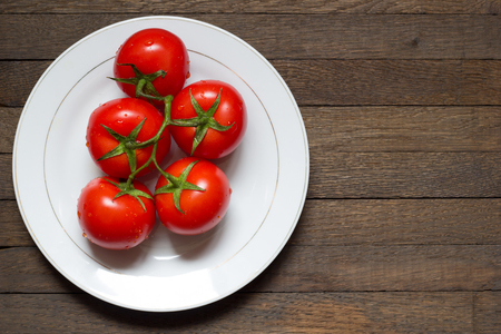 Red ripened tomatoes on white plate on wooden table with copy space. Raw foods. Healthy nutrition. Washed vegetables on branch with water droplets on surface. Close up. Top view. Left position.の写真素材