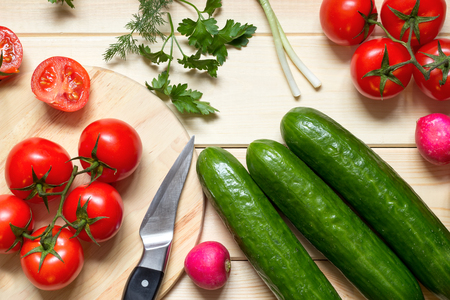 Cooking food from vegetables and greens. Healthy nutrition concept. Fresh raw ingredients: cherry tomatoes, cucumbers, radish, parsley, dill, green onion. Knife on kitchen board. Top view. Close-up.の写真素材