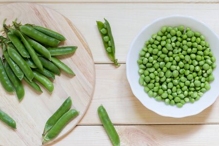 Healthy organic natural home-grown vegetables. Green sugar snap peas in white plate and on kitchen board on light wooden table. Whole pods and peeled beans. Top view background. Close-up.の写真素材