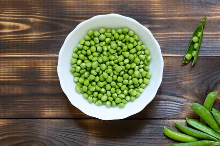 Healthy eating concept. Fresh green peas in white plate on dark wooden table. Whole pods and one open pod. Copy space. Place for text. Burned texture. Top view. Close-up. Natural organic products.の写真素材