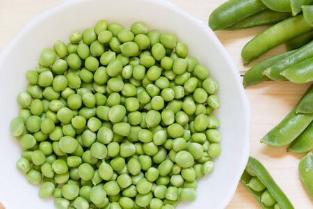Fresh green peas in white plate on wooden table. Peeled delicious vegetables and whole pods lay on light wooden table. Healthy nutrition concept. Top view. Close-up. Horizontal photography.の写真素材
