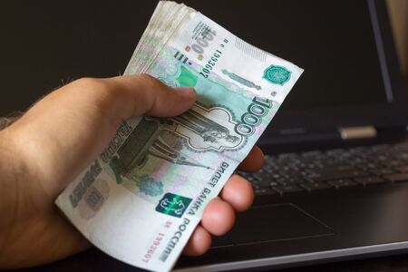 Man hand holding bundle of Russian money. Laptop in background.