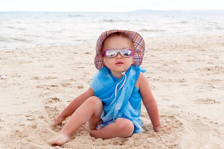 A little girl in blue pareo, sunglasses and a hat sits on the golden sand on the beach by the sea. Tanned skin with slight irritation. Vacation with children near water.の写真素材