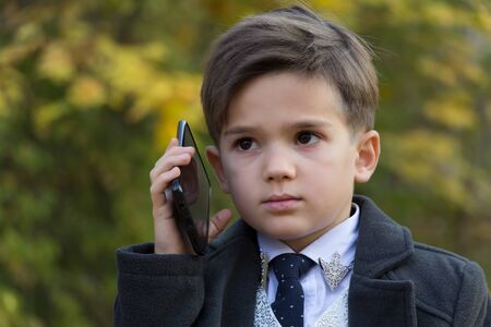 Portrait of pensive little businessman talking by phone on autumn background. The development of modern children concept. Little adults. A small man dressed in a business suit, white shirt and tieの写真素材