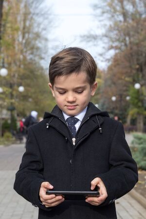A schoolboy enthusiastically clicks on the screen of his smartphone, holding it in both hands in a horizontal orientation. Front view. Autumn alley. Selective focus on a boy in a business coatの写真素材