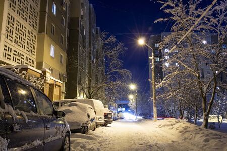 Snow-covered city courtyard in the late evening. Magical landscape. Snow-covered cars parked along high-rise buildings, lights of lanterns. Winters Tale in Donetsk, Ukraine. Scenic view from roadの写真素材