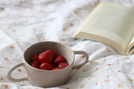Unrecognizable person holding a bowl of strawberries. Open book on floral sheets. Selective focus.の写真素材
