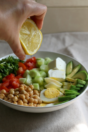 Squeezing lemon juice on lunch bowl with avocado, hard boiled egg, chickpea, feta cheese, cucumber, tomato and arugula. Selective focus.の写真素材