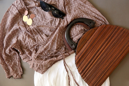 Fashionable outfit with blue nude dotted blouse, white skirt, vintage wooden bag, sunglasses and gold earrings. Flat lay, gray background.の写真素材