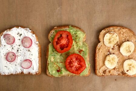 Three different toasts: cream cheese with radish and basil, avocado with cherry tomato and smoked paprika, peanut butter with banana and cinnamon. Top view, brown background.の写真素材
