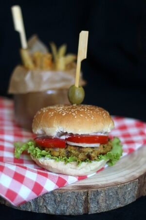 Homemade vegan burger made with chickpeas and zucchini, served with lettuce, tomato, cucumber, pickles and sauce. Fries on the side. Selective focus, dark background.の写真素材