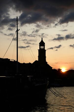 Beautiful sunset in small town Sutivan, on island Brac, Croatia. Silhouettes of historical bell tower, traditional homes and small boats against the colorful cloudy sky.の写真素材