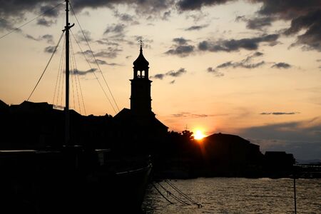 Beautiful sunset in small town Sutivan, on island Brac, Croatia. Silhouettes of historical bell tower, traditional homes and small boats against the colorful cloudy sky.の写真素材