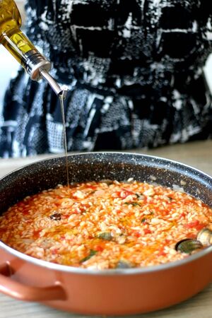 Unrecognizable person pouring olive oil in pot of orzo pasta, with tomato sauce, vegetables and seafood. Selective focus.の写真素材