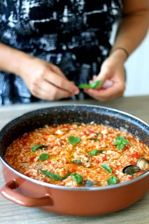 Unrecognizable person putting basil leaves in pot of orzo pasta, with tomato sauce, vegetables and seafood. Selective focus.の写真素材
