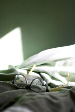 Grey and green reading glasses on a bed with green sheets and green wall. Selective focus.の写真素材