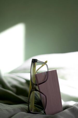 Book and reading glasses on a bed with green sheets, illuminated by sunlight. Selective focus.の写真素材