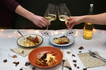 Festive holiday table with pork belly roast, mashed potatoes and roasted vegetables. Unrecognizable people cheering with white wine.の写真素材