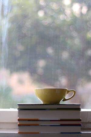 Tea in a yellow cup and stack of books on a rustic window sill. Selective focus, view of garden in the background.の写真素材